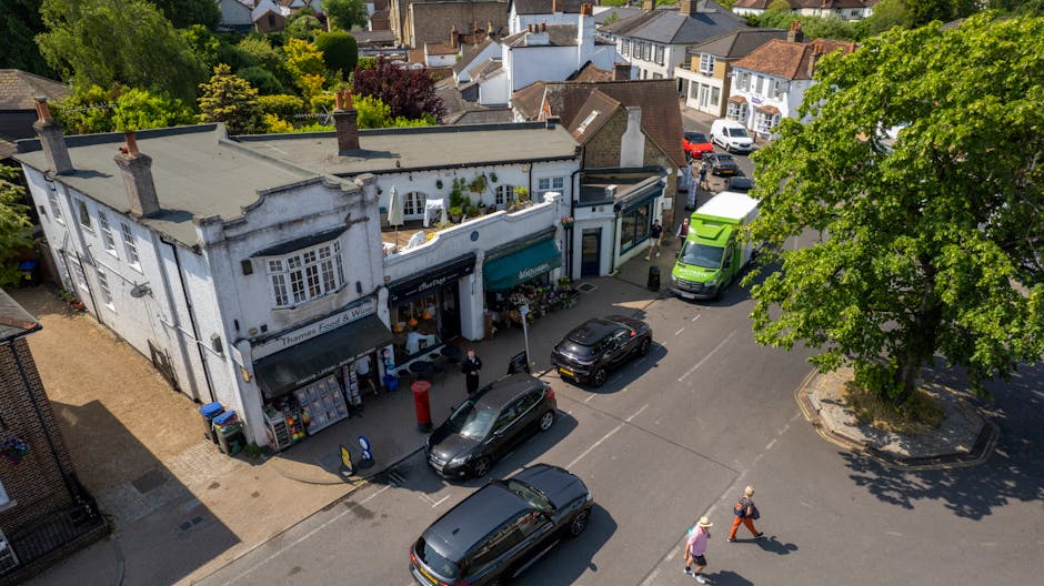 An aerial view of a small urban street in Hampton Hill with a white building featuring a shopfront and an awning, surrounded by parked cars and delivery vans. The scene includes a large green van and a black car positioned near the building, with two pedestrians walking on the pavement. A person appears to be loading or unloading items from a van close to the shop, indicating a home relocation or moving process. The street is lined with trees and residential houses in the background, with some greenery visible. The overall environment is well-lit with natural sunlight, capturing the typical moving logistics scenario involving furniture and boxes being transported in a busy neighbourhood, reflecting the service capabilities of Man With a Van Hampton Hill for address access solutions during house removals.