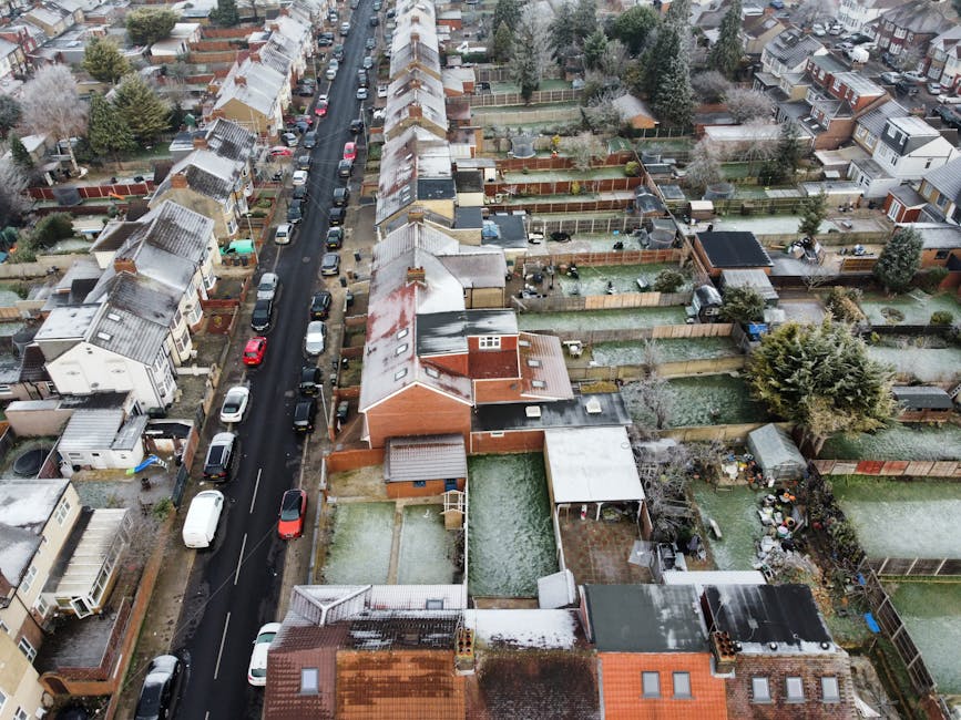 An aerial view of a residential street in Hampton Hill showing closely spaced terraced houses with pitched roofs, some of which have a light dusting of snow. Cars are parked along both sides of the narrow street, and the street is lined with trees and lampposts. Behind the houses, there are fenced gardens with various outdoor furnishings, sheds, and garden equipment. The scene depicts a typical urban setting with a mix of brick and weatherboard exteriors, narrow alleyways, and backyards. The image highlights the challenges of accessing homes on narrow streets for removals, and is relevant to home relocation or furniture transport services such as those provided by Man With a Van Hampton Hill, especially dealing with access problems on narrow Hampton Hill streets and planning efficient loading processes for house removals.