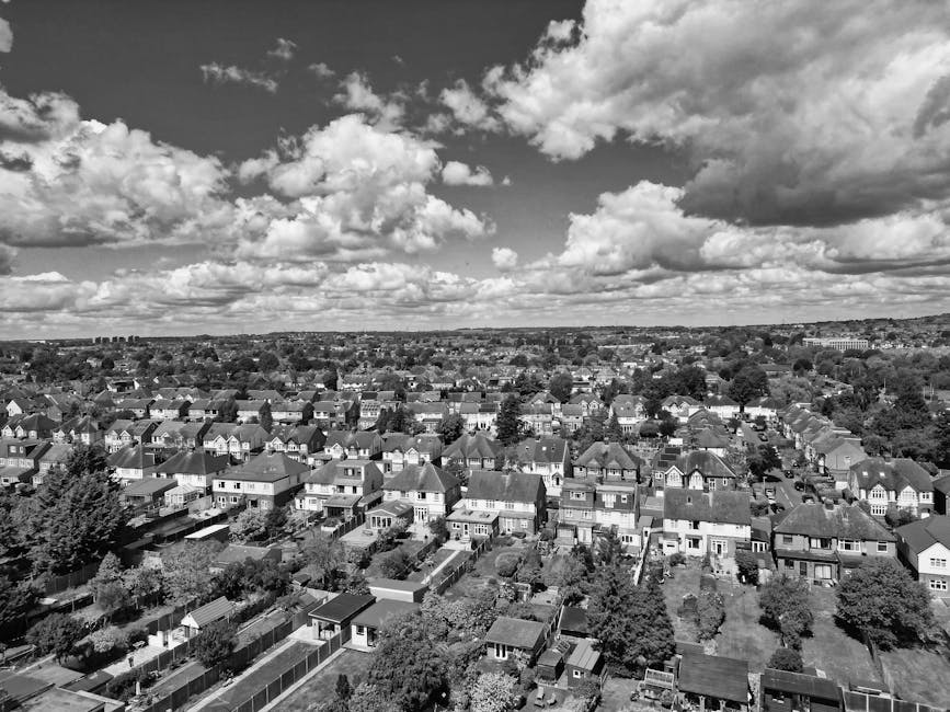 A high-angle black and white photograph of a suburban residential area showing numerous houses with pitched roofs, some with chimneys, arranged in a grid-like pattern with small gardens and backyards enclosed by fences. The streets run parallel and perpendicular, lined with trees and parked cars. In the foreground, there are smaller outbuildings and sheds within backyards. The sky above is filled with large, scattered clouds adding contrast to the scene, with no movement visible. This image captures a quiet, well-established neighbourhood suitable for home relocation and furniture transport, reflecting the types of residential environments serviced by Man With a Van Hampton Hill. The overall scene emphasizes the typical layout of London suburbs, highlighting the importance of proper packing and loading during a house removal.