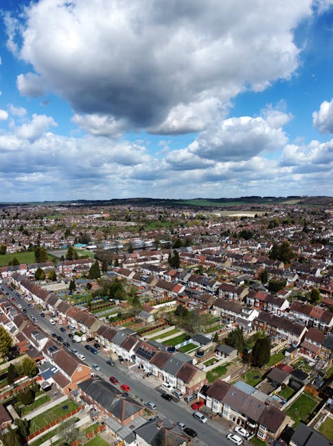An aerial view of a residential neighbourhood under a partly cloudy sky, showing rows of terraced houses with red and brown tiled roofs, small front gardens, and parked cars along the streets. Some houses have driveways and green lawns, with trees interspersed between properties. In the foreground, an area of pavement and parked vehicles suggests a loading zone commonly used during home relocation or furniture transport. The image captures the extensive layout of the suburban area, illustrating the environment in which house removals and packing and moving activities by companies like Man With a Van Hampton Hill typically take place. The natural lighting and open sky create a clear scene of the locality, emphasizing the logistical considerations involved in local removals services within the Hampton Hill area.