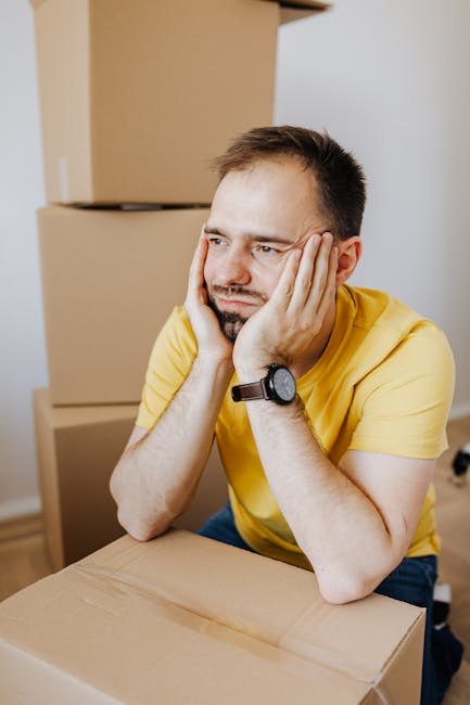 A man wearing a yellow t-shirt and a black wristwatch is sitting at a table in a room filled with cardboard boxes. He rests his head on his hands with a distressed expression, suggesting frustration or worry. Behind him, stacked cardboard boxes and a partially visible doorway indicate a home ready for packing or recent packing activities. The scene captures a moment of pause during house removals or packing preparations, with the boxes and indoor environment highlighting the ongoing relocation process that Man With a Van Hampton Hill manages as part of their removals services.