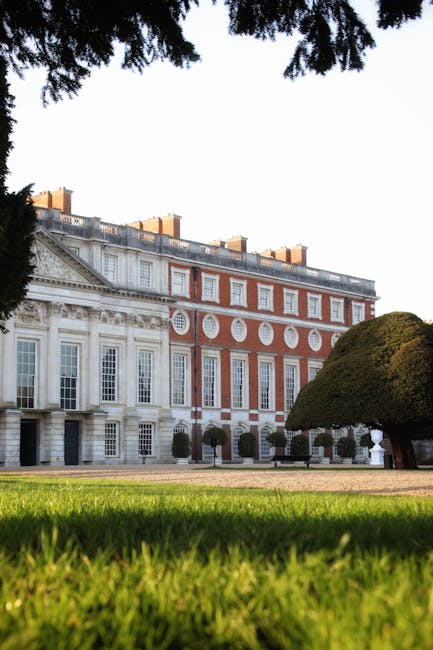 A large, historic building with a white and red brick facade, featuring tall sash windows and decorative architectural details, situated behind a well-maintained lawn and landscaped garden with trimmed trees. The scene is captured from ground level, with the foreground of green grass and a partial view of a large, rounded tree on the right. Overhanging tree branches frame the top of the image, and the lighting suggests daylight with a clear sky. This setting could be associated with the scenic route for house removals or home relocation services between Bushy Park and Hampton Court, as offered by Man With a Van Hampton Hill, showing an example of a notable landmark or destination in the area.
