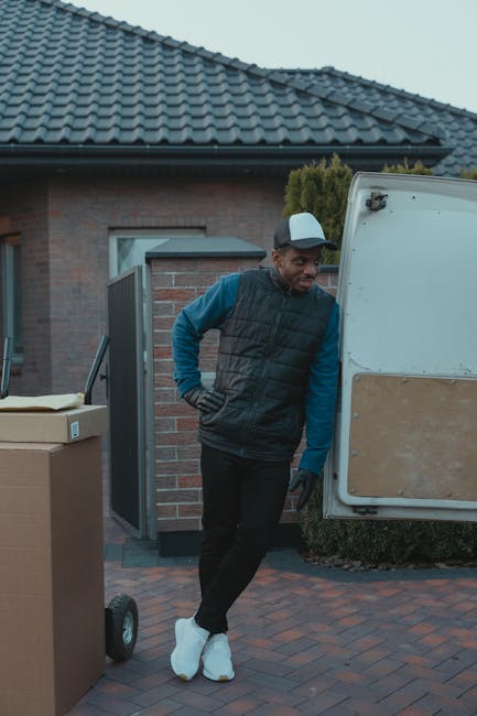 A man wearing casual clothing and a cap is seen carrying three cardboard boxes of varying sizes, stacked and held together with his arms, during a home relocation process. He is walking on a suburban 