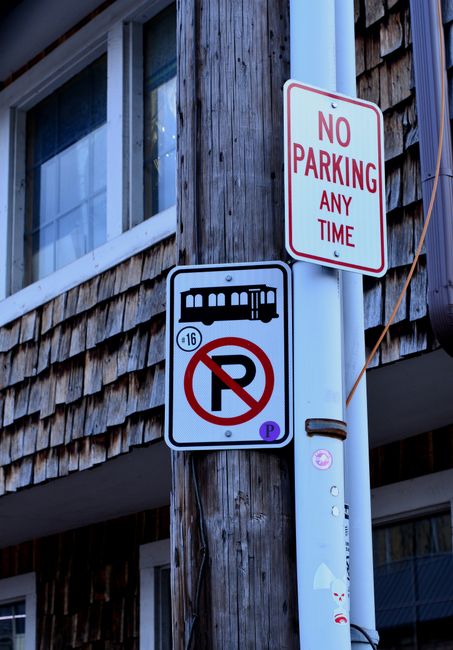 Close-up view of a wooden building exterior with two traffic signs mounted on a metal pole. The top sign is a rectangular 'No Parking Any Time' sign with red text on a white background, affixed to the