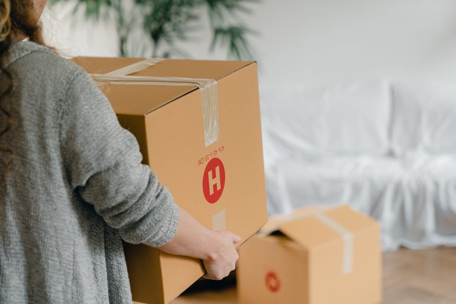 A person wearing a grey knitted sweater is in the process of packing a large cardboard box with moving supplies inside a home. The box is sealed with clear packing tape, and the individual is holding a utility knife, applying it to the tape to secure the contents. In the background, additional cardboard boxes, also sealed with tape, are positioned on a wooden floor near a white sofa with draped cushions. Natural light illuminates the scene, highlighting the details of the furniture and packing materials, which include the cardboard boxes and tape used for secure packing. This image exemplifies the packing and furniture transport aspect of home relocation services, as provided by Man With a Van Hampton Hill, emphasizing careful handling and organized packing during moving and removals within a residential setting.