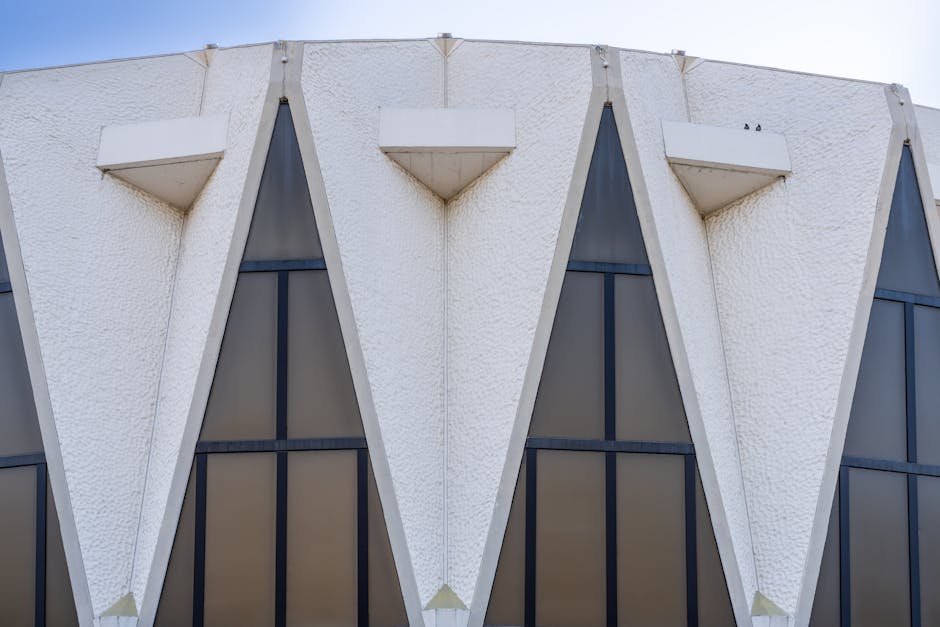 Close-up view of a modern building's exterior facade featuring a series of large, vertically oriented, triangular glass windows framed by white textured concrete with angular projections. The building's design includes three prominent triangular window sections with dark-tinted glass, separated by bold white concrete supports that extend outward, creating a repetitive geometric pattern. Small white protruding ledges are positioned above each window section, and the overall structure is illuminated by natural daylight, highlighting the clean lines and architectural style. The image captures the detailed surface texture and material contrast, reflecting contemporary architectural elements related to building design and construction, which may be relevant to home relocation or renovation contexts managed by Man With a Van Hampton Hill.