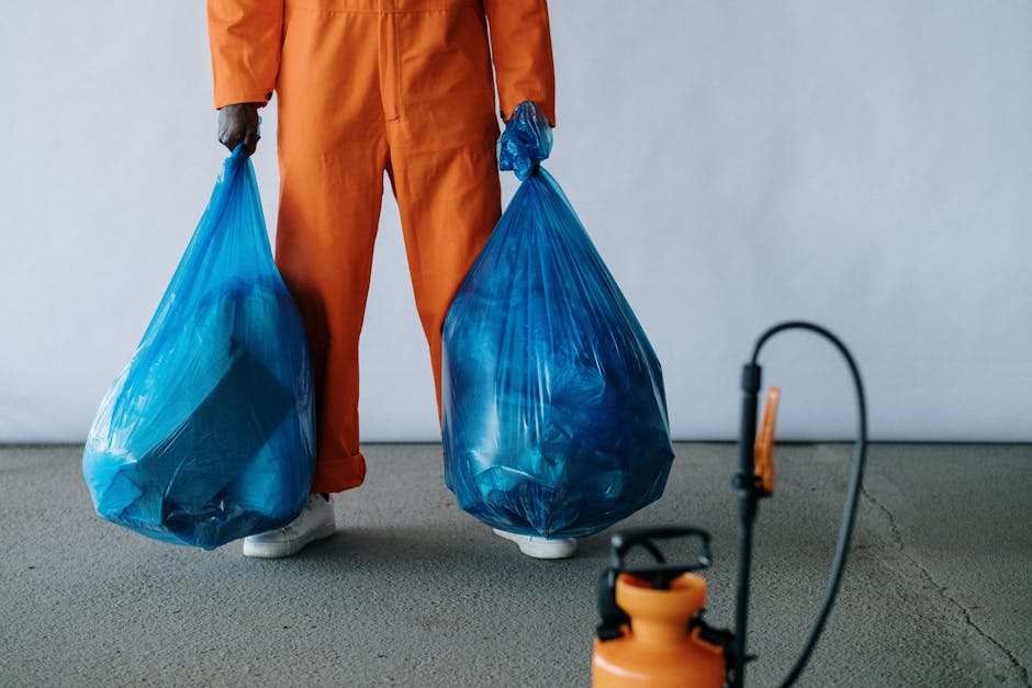 A person dressed in an orange jumpsuit and white shoes is standing indoors, holding two large blue plastic rubbish bags filled with waste. The person's face is not visible, and they are positioned with their legs apart on a plain gray floor against a light-colored wall. To the right, partially in view, is a black and orange handheld sprayer or cleaning device, connected by a flexible hose. The scene appears related to house removals and packing procedures, with the waste bags likely collected during a home relocation process. The setting suggests a preparation or cleaning stage prior to or during a move, with focus on waste disposal consistent with local rules for waste during house removals, as indicated by the page title from Man With a Van Hampton Hill on their website.