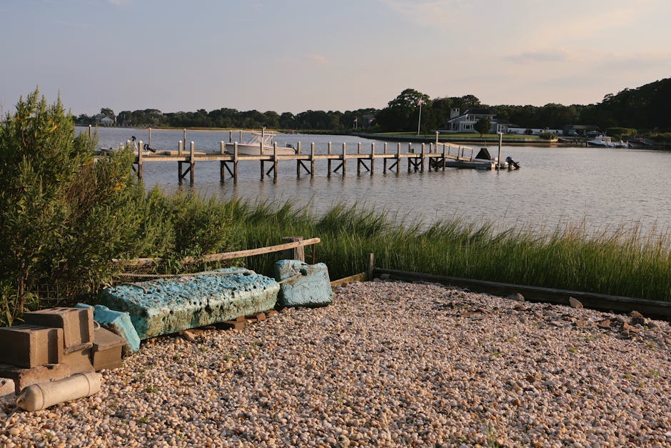 A gravelly outdoor area adjacent to a body of water with a dock extending into the river, featuring a pile of weathered blue foam insulation boards, some wooden planks, and a grey plastic pipe lying on the ground. Behind the foam boards, there are green shrubs and tall grass, with a metal fence or barrier on the right edge. The water appears calm, with several boats moored along a shoreline that has residential buildings and trees in the background. The scene is illuminated by natural daylight, suggesting a clear or partly cloudy sky. This setting could be associated with home relocation or the loading process, as part of furniture and packing materials are visible near the waterfront, potentially prepared for transportation by Man With a Van Hampton Hill during a house move or furniture transport. The overall scene captures an environment conducive to moving logistics, including outdoor storage or staging for a house removal service.