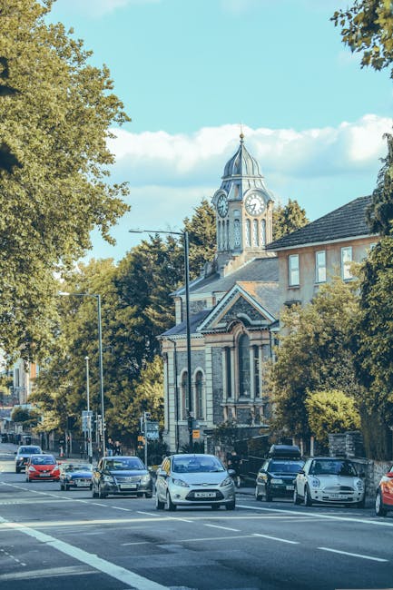 A busy street scene in Hampton Hill showing a row of parked cars along the pavement, with various models and colours, including white, black, and silver vehicles. In the background, there is a historic church building with a distinctive clock tower topped by a pointed roof and a weather vane, partially surrounded by large leafy trees providing shade and greenery. The sky is bright blue with some scattered white clouds, and the street is illuminated by natural daylight. Street lamps and traffic signs are visible along the roadside, and the scene suggests a typical urban environment suitable for house removals and furniture transport services, as undertaken by Man With a Van Hampton Hill. This setting highlights the importance of careful logistical planning during home relocation or packing and moving processes in the area.