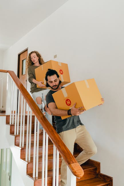 A man and a woman are carrying cardboard boxes during a home relocation inside a house. The man, positioned on the staircase, is holding a medium-sized cardboard box with packing tape sealing it, leaning forward as he descends the wooden stairs. The woman, standing behind him on the stairs, is holding a larger box and appears to be guiding or assisting with the move. Both are dressed casually, and the staircase has a wooden handrail and white railings, with a light-colored wall and a door visible at the top. This scene depicts the process of packing and furniture transport as part of a professional removal or moving service, with an emphasis on the careful handling of boxes during a house move, consistent with the services offered by Man With a Van Hampton Hill.
