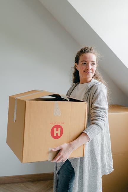 A woman with curly hair wearing a light grey sweater carries a brown cardboard box with the red 'H' logo of Man With a Van Hampton Hill, inside a room with a sloped ceiling and white walls. Several other similarly packed boxes are visible in the background, positioned on the wooden floor near the wall. The woman appears to be involved in a home relocation process, preparing for furniture transport or packing and moving activities. The image captures the loading process in a residential setting, with natural lighting illuminating the scene, highlighting the careful handling of packing materials during the house removal operation.