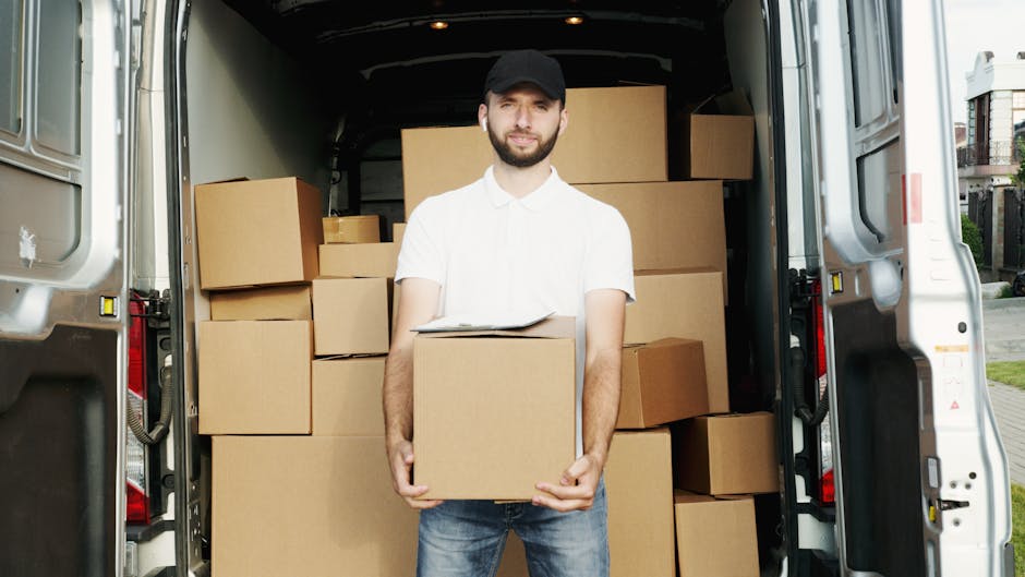 A man with a beard, wearing a white polo shirt and a black cap, stands in the open rear of a commercial van loaded with cardboard moving boxes of various sizes. The boxes are neatly stacked inside the van, some on the floor and others on top, secured for safe transport. The loading area is well-lit with natural daylight, showing a residential street in the background with houses and greenery. The man holds a medium-sized cardboard box in front of him and appears ready for the home relocation process, representing furniture transport and packing during a house move. The scene reflects the loading process involved in professional removals, with a focus on secure packing and careful handling, consistent with services offered by Man With a Van Hampton Hill.