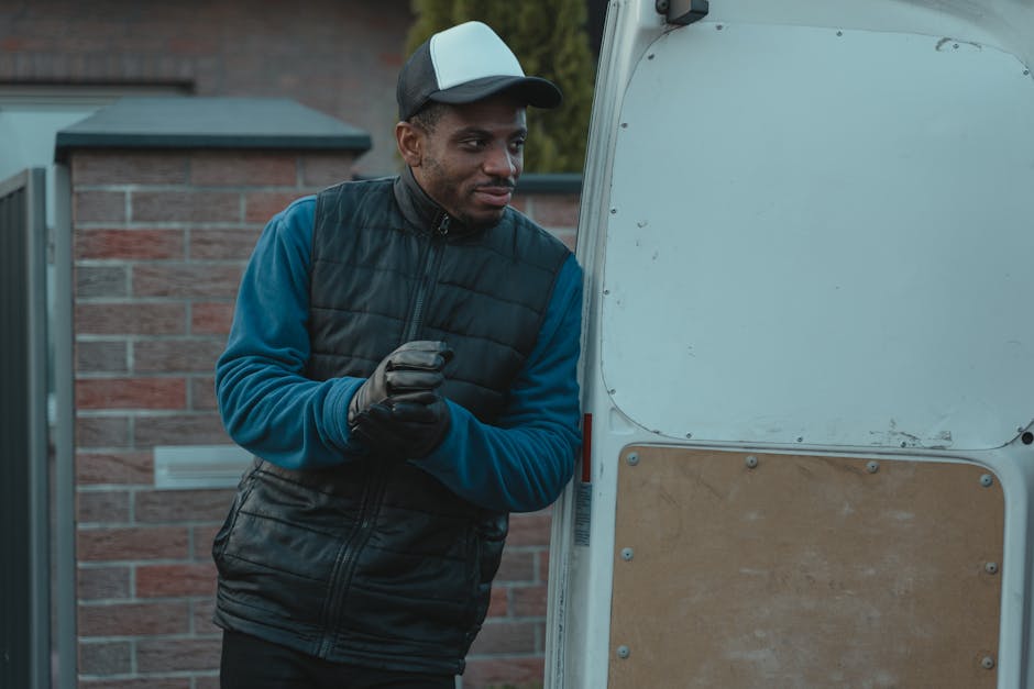 A man wearing a dark baseball cap, a blue long-sleeve shirt, and a black padded vest, stands outdoors near the back of a white moving van with a partially open cargo door. He is holding a pair of gloves and appears to be preparing for a home relocation or furniture transport. Inside the van, cardboard moving boxes and wrapped furniture pieces are visible, indicating packing and moving preparations. The setting is an outdoor driveway with a brick wall and a gate in the background, under natural daylight. The man with a van company, Man With a Van Hampton Hill, is engaged in the loading process, which involves arranging and possibly lifting items into the vehicle for a house removal or local move, highlighting the logistics involved in packing, securing, and transporting household belongings.