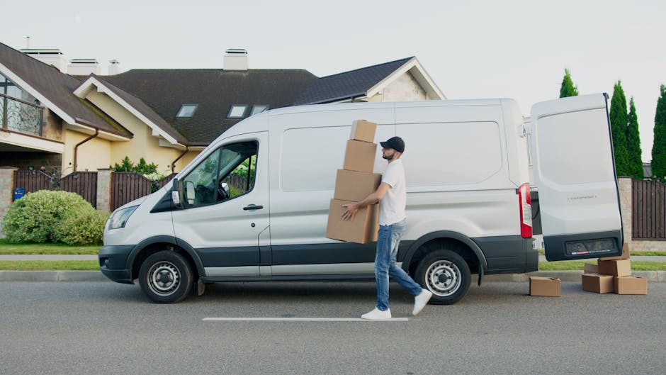 A man with a beard, wearing a white polo shirt and a black cap, stands in the open rear of a commercial van loaded with cardboard moving boxes of various sizes. The boxes are neatly stacked inside the van, some on the floor and others on top, secured for safe transport. The loading area is well-lit with natural daylight, showing a residential street in the background with houses and greenery. The man holds a medium-sized cardboard box in front of him and appears ready for the home relocation process, representing furniture transport and packing during a house move. The scene reflects the loading process involved in professional removals, with a focus on secure packing and careful handling, consistent with services offered by Man With a Van Hampton Hill.